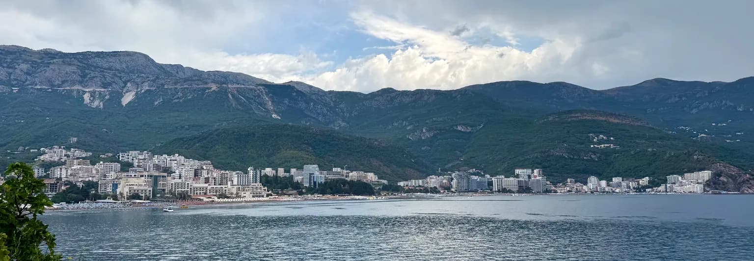Plaża Bečići i nadmorskie hotele na Riwierze Budvańskiej, widziane z drugiej strony zatoki, z górami w tle.